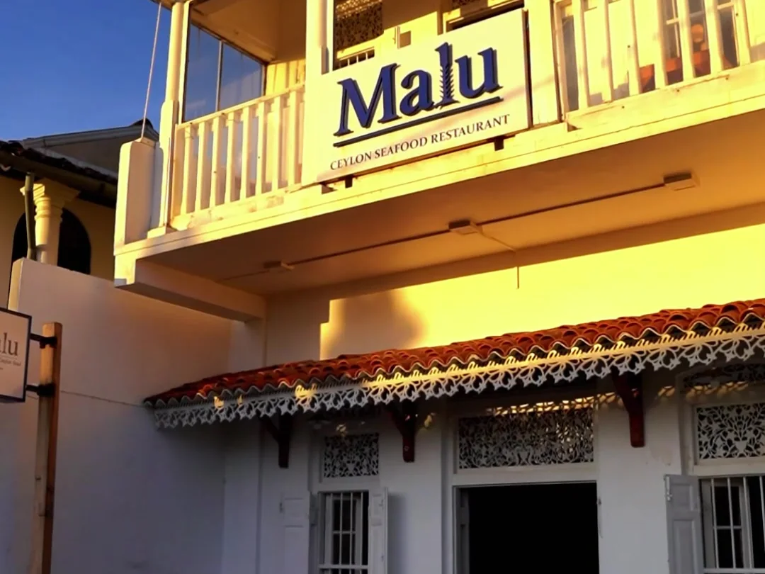 Fachada de Malu con luz dorada: balcón amarillo con el rótulo «Malu, Ceylon Seafood Restaurant» y la entrada de la villa